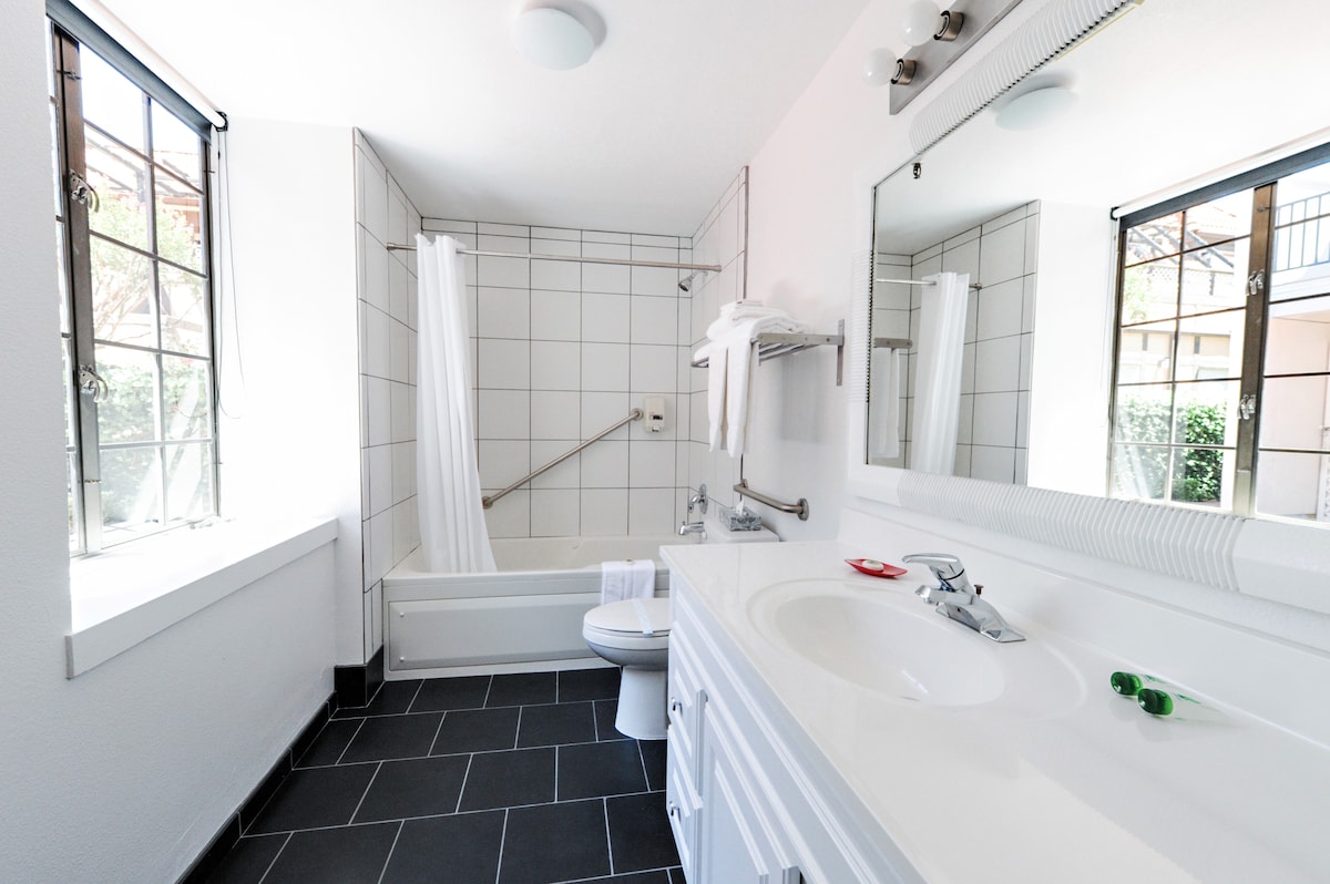 The bathroom features a tub and shower combination with a safety grab bar. A modern sink is positioned beside a large mirror. Natural light enters through a window, while white towels are neatly arranged on a nearby rack.