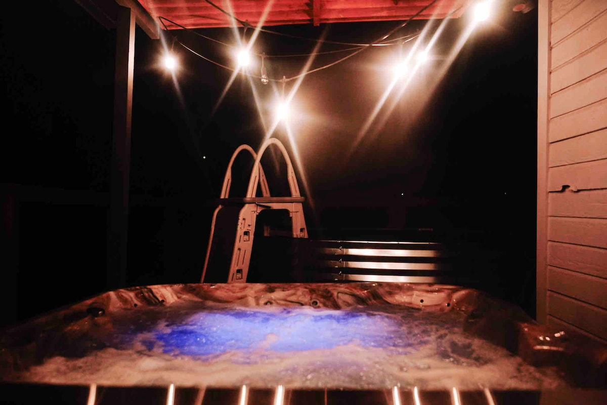 The hot tub is illuminated by soft glowing string lights, creating a cozy atmosphere. A safety ladder is positioned nearby. The surface of the water reflects a calming blue hue, inviting relaxation beneath the night sky.