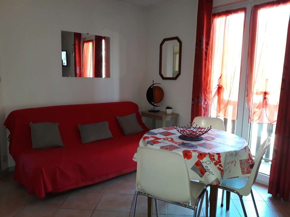 A cozy living area is shown with a red sofa bed complemented by several gray cushions. A round dining table with a patterned tablecloth is surrounded by four white chairs. Bright curtains frame the windows, and a decorative globe is positioned on a nearby table.