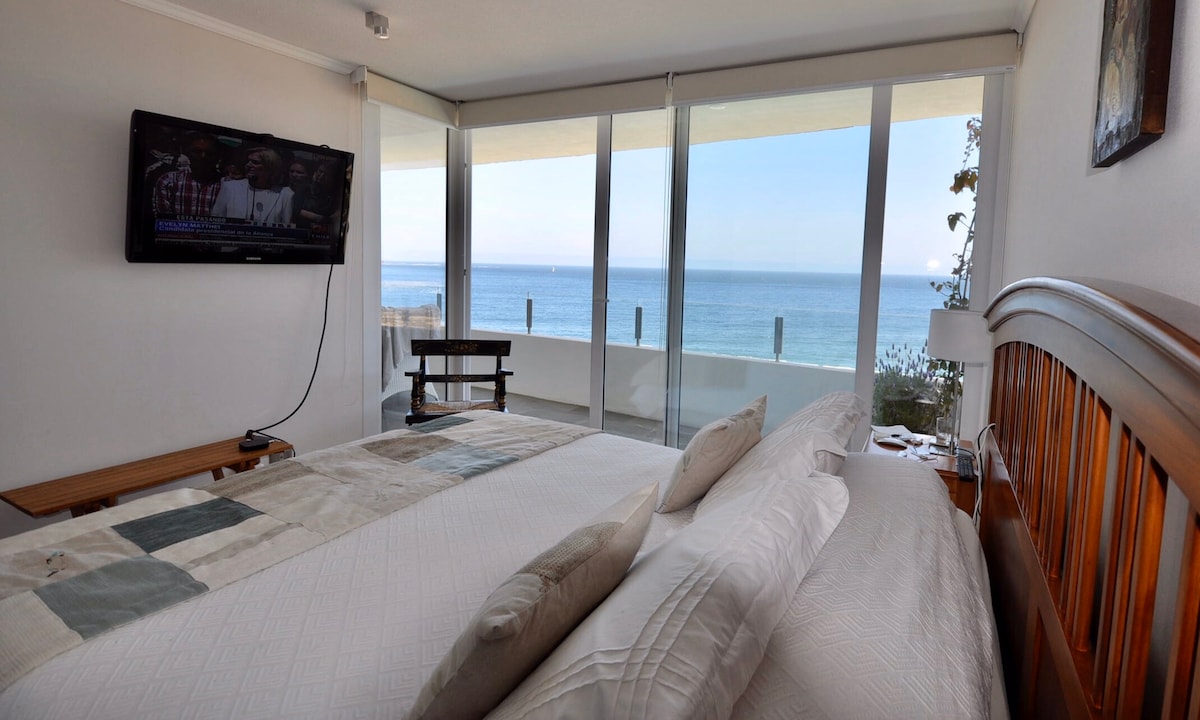 A bright, airy bedroom features a spacious bed adorned with neutral-toned linens. Large glass doors provide direct views of the ocean, enhancing the room's light and openness. A flat-screen television is mounted on the wall, and a small table is positioned beside the bed.
