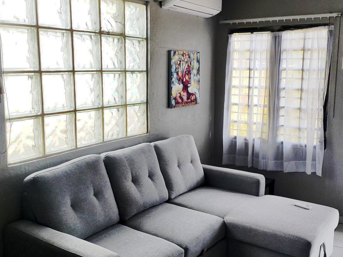 A gray sectional sofa is positioned near a large window, allowing natural light to fill the room. A colorful artwork hangs above the sofa, while sheer curtains frame the window, creating a bright and airy atmosphere.