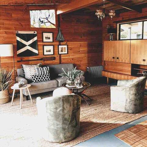 A living area is presented with timber paneling that creates a warm atmosphere. A gray couch is complemented by two circular chairs and a glass coffee table adorned with a plant. Artwork is displayed on the walls, and light enters through the large window.