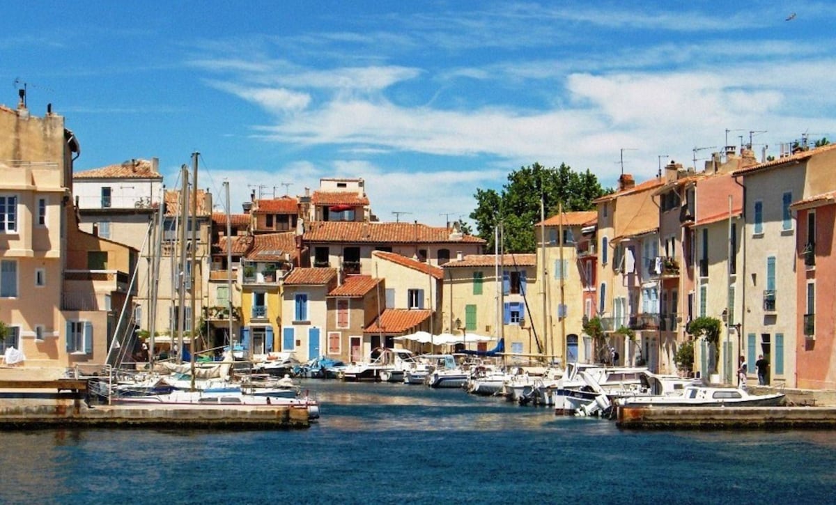 A charming waterfront scene captures colorful buildings lining a canal, complemented by various boats docked along the shore. Clear blue skies reflect on the water, and lush greenery is visible in the distance, creating a vibrant and serene environment.