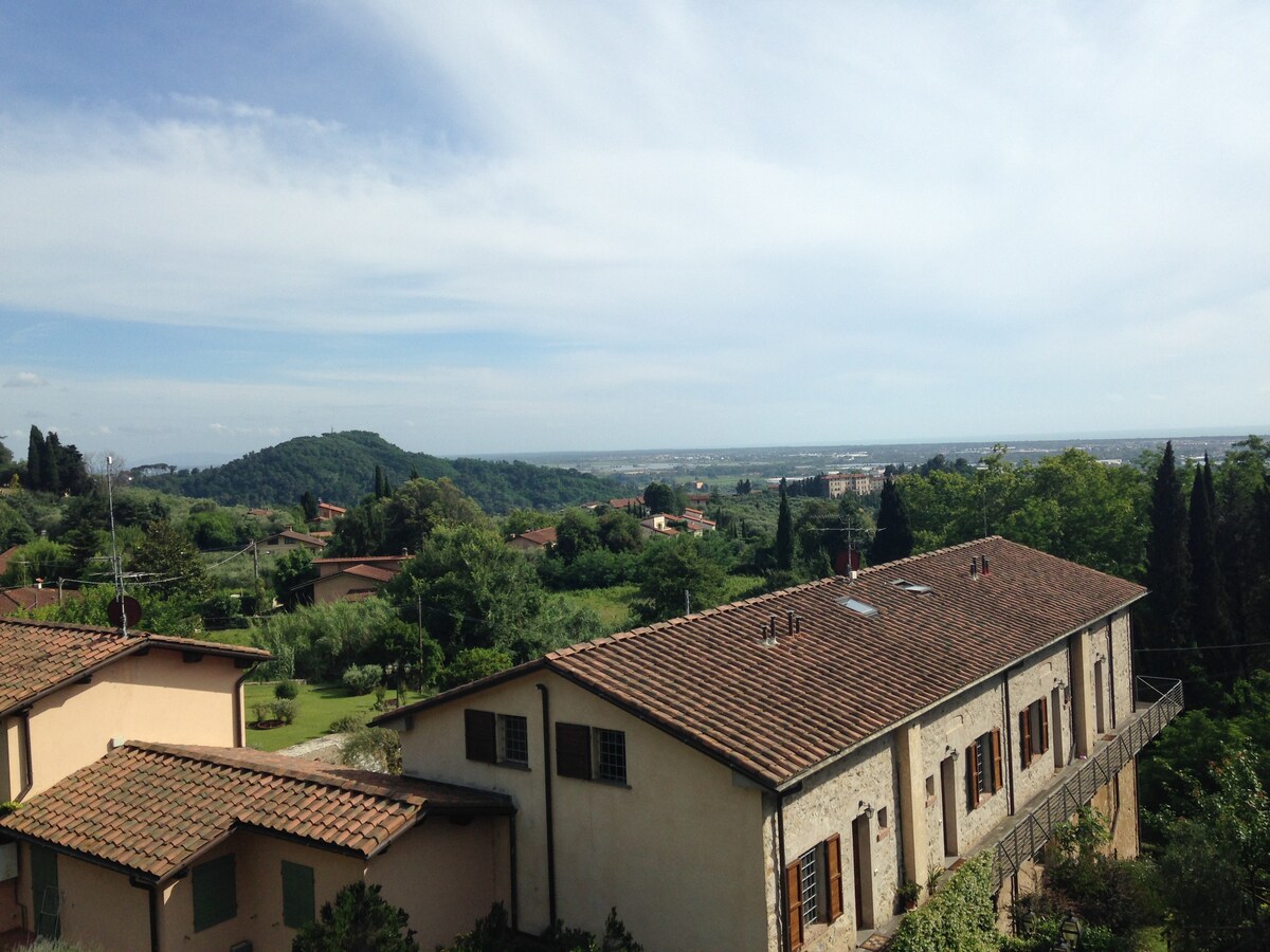A panoramic view showcases the rural landscape, with rolling hills and lush greenery in the foreground leading to a distant horizon. The low-rise building features a terracotta-tiled roof, and the surroundings include various trees and vegetation, typical of a serene countryside setting.