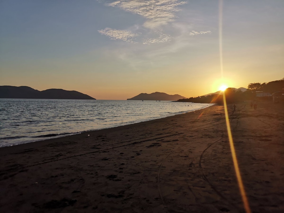 The image captures a serene beach at sunset, with soft waves lapping against the shore. Distant hills are silhouetted against the warm hues of the sky, where the sun is slowly descending. Soft footprints and tire tracks mark the sandy beach, suggesting a tranquil environment.