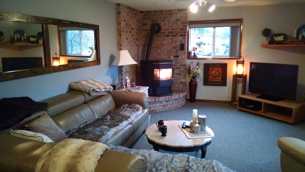 A cozy living room is highlighted by a warm, inviting fireplace set within a brick surround. A comfortable sectional sofa is adorned with soft throws, while a round coffee table sits in the center. Natural light enters through a large window, enhancing the space's comfort.