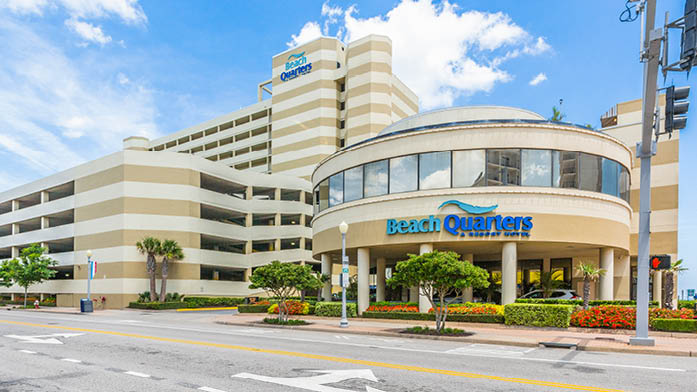 The exterior of Beach Quarters Resort is showcased, featuring a modern design with cream and beige striped walls. A round, glass entrance highlights the main lobby area, surrounded by lush green landscaping. A multi-level parking garage is visible in the background, under a clear blue sky.