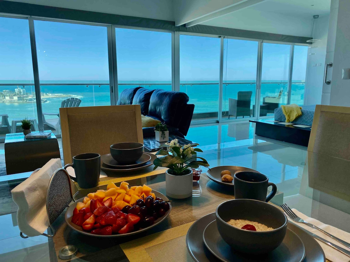 An inviting dining table is set with bowls of fruit, yogurt, and coffee, arranged alongside a small potted plant. Through floor-to-ceiling windows, a panoramic view of the ocean and distant coastline is visible, creating a serene backdrop for a meal.