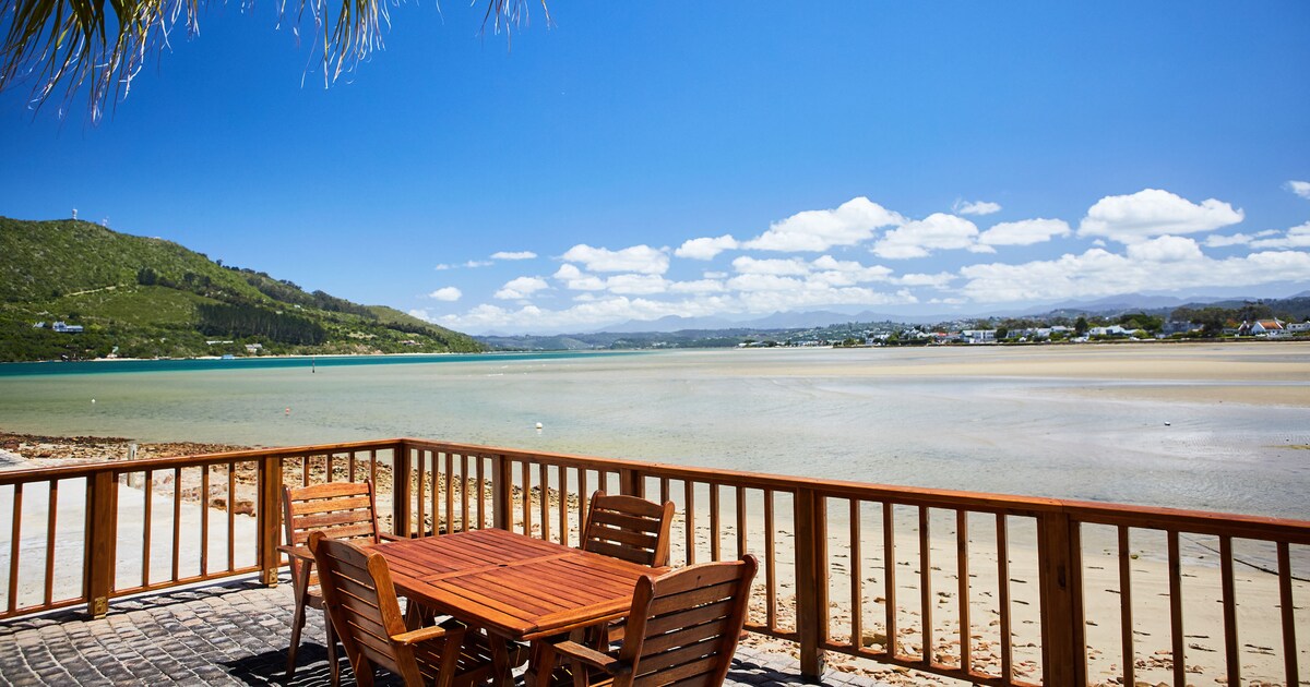 A wooden deck features a table and chairs, providing a space for outdoor dining with expansive views of the lagoon and distant hills. The clear blue sky and soft clouds create a serene backdrop, emphasizing the tranquil setting of the waterfront.