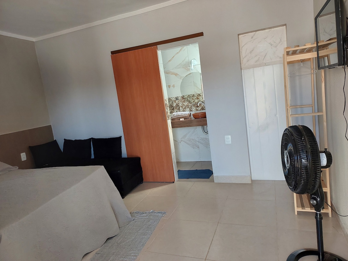 A bright and airy room is showcased, featuring a spacious bed with a light cover and a soft rug on the tiled floor. A small seating area is visible with dark cushions. A door leads to a bathroom, and a freestanding fan adds comfort.