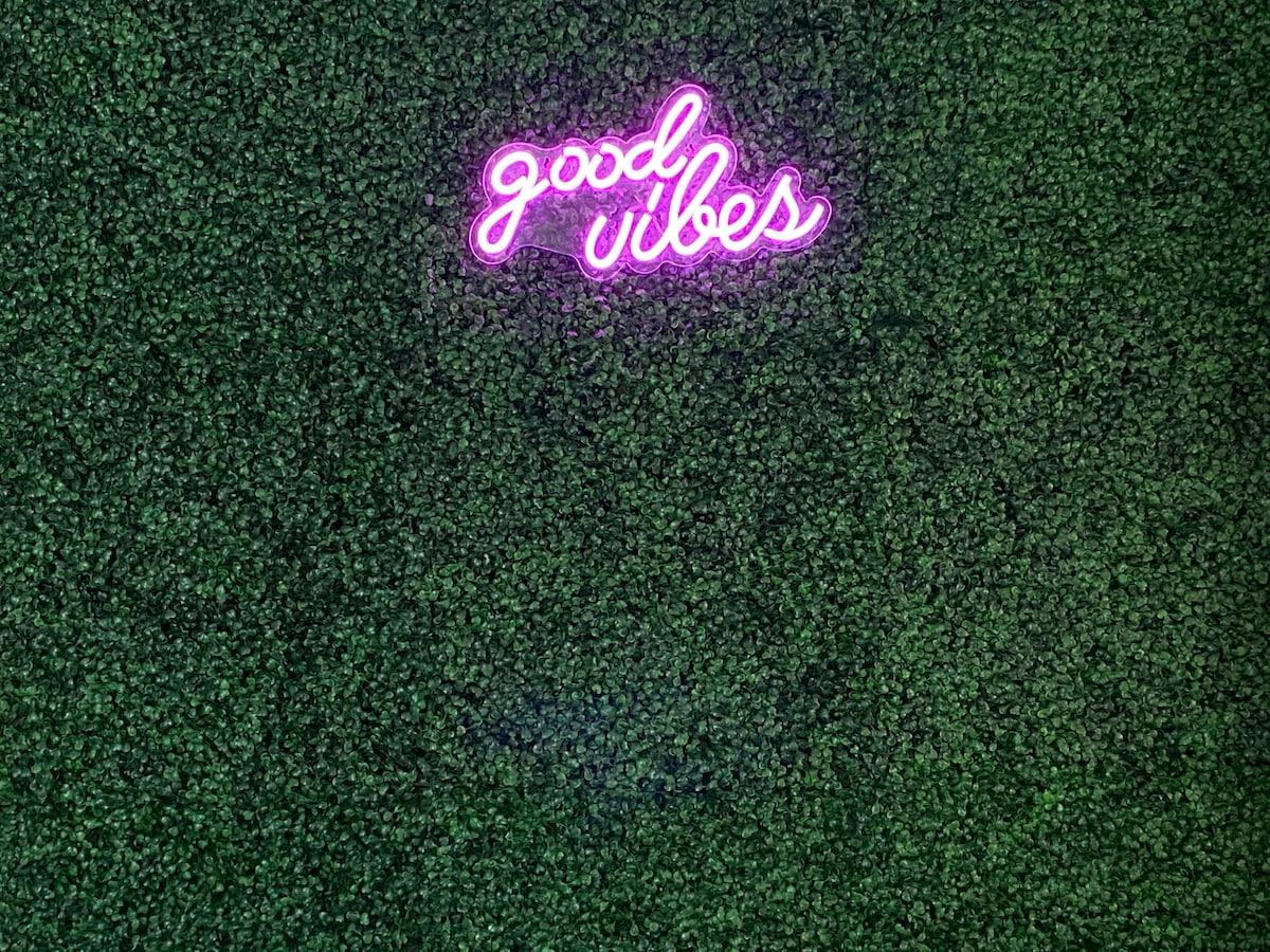 A vibrant neon sign displaying the words 'good vibes' is mounted against a textured green wall, creating a lively focal point. The wall features a lush appearance, enhancing the playful atmosphere of the space.