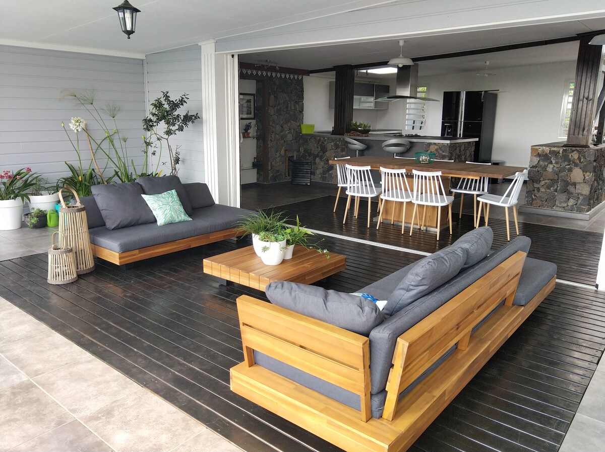 An open-air living space features comfortable seating arrangements with wooden furniture. A coffee table sits at the center, surrounded by potted plants. The backdrop includes a dining area furnished with white chairs and a stone wall, enhancing the overall spaciousness.