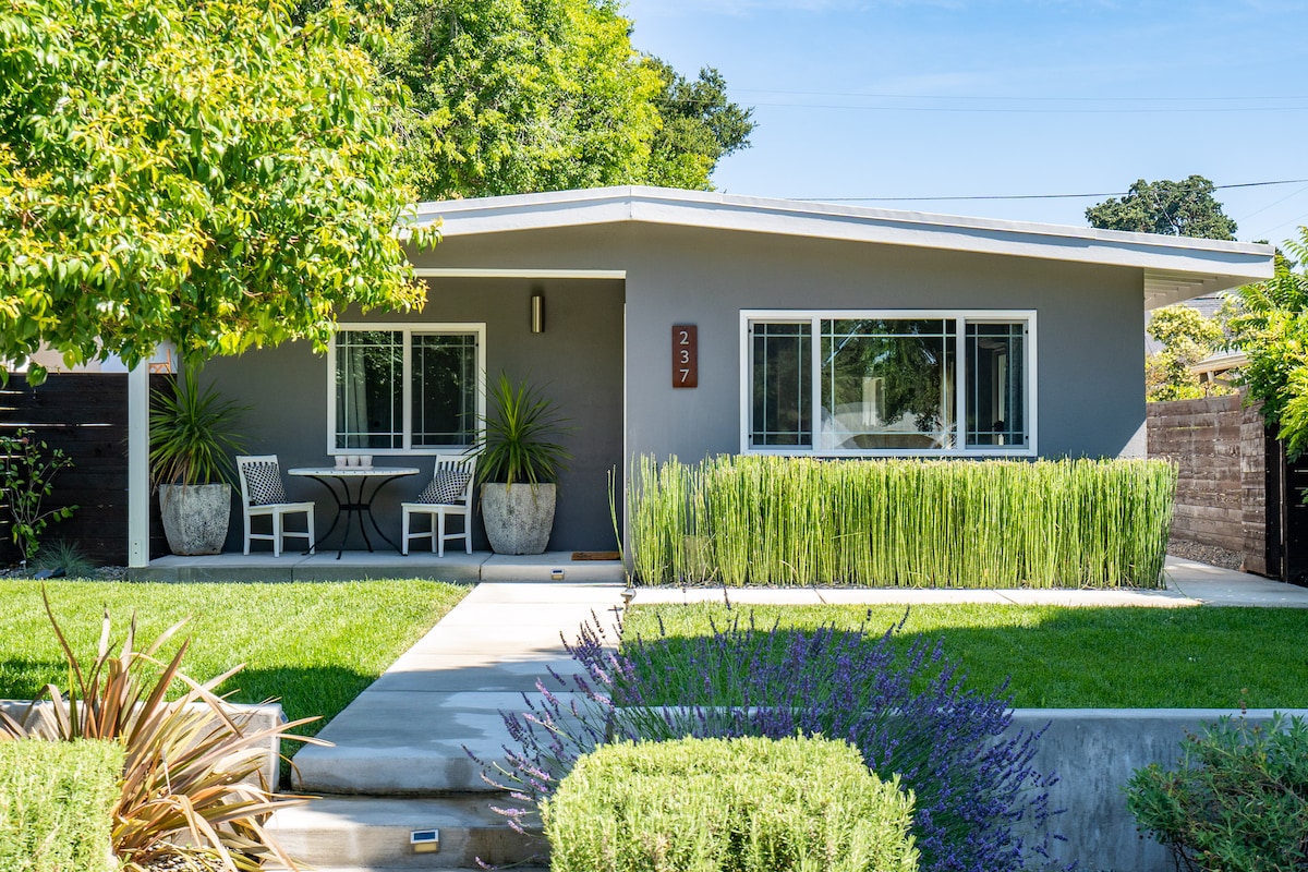 A Mid-Century Modern home is set amidst lush landscaping, featuring a symmetrical front layout. Potted plants accentuate the entrance, while a small outdoor seating area is visible near the front porch. The manicured grass and vibrant greenery contribute to a welcoming ambiance.