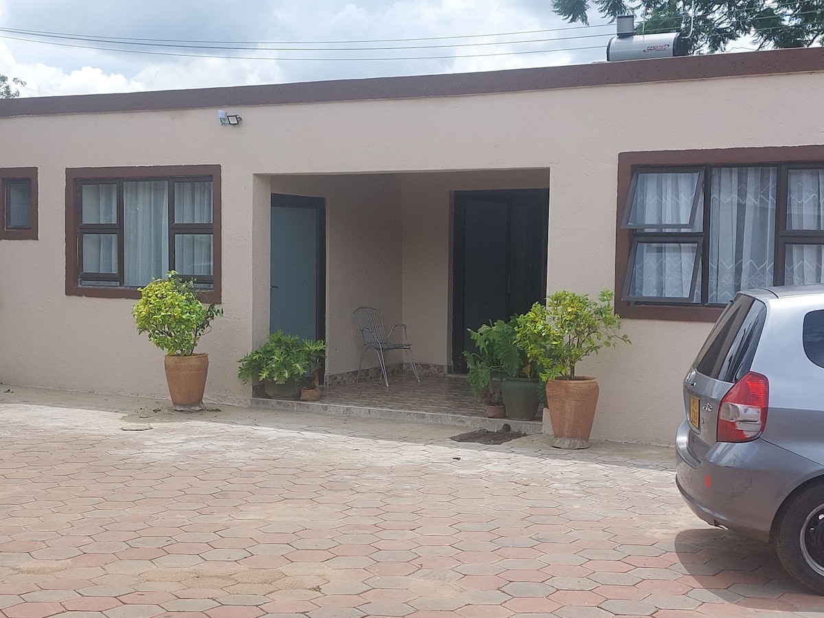 An exterior view of a single-story building is presented, featuring a covered entryway. Potted plants are arranged on either side of the entrance, providing greenery. A gray vehicle is parked nearby on a paved surface, under a partly cloudy sky.