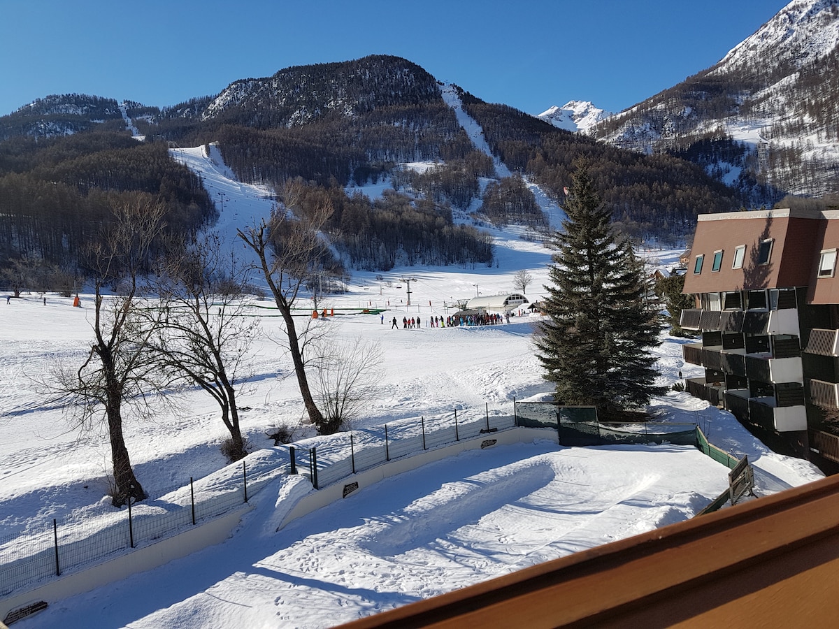 A winter landscape showcases a snowy slope with skiers in the distance, surrounded by majestic mountains. Several trees with bare branches are visible in the foreground, while a building with balconies stands nearby. The clear blue sky enhances the overall serene setting.