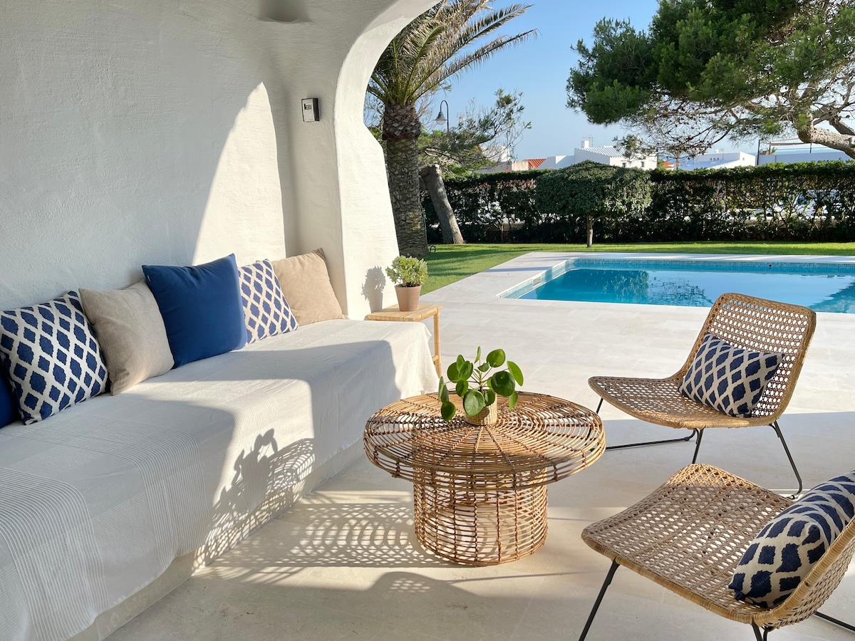 A covered seating area is presented with a spacious white couch adorned with patterned cushions. A rattan coffee table occupies the space alongside two woven chairs. In the background, the private pool glimmers under the sun, bordered by well-maintained greenery.