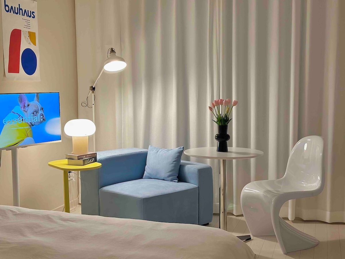 A cozy seating area features a light blue couch next to a small yellow table. A modern white chair and a vase with pink flowers sit on the white floor. A flat-screen TV is mounted on the wall, complemented by a stylish floor lamp, against soft white curtains.