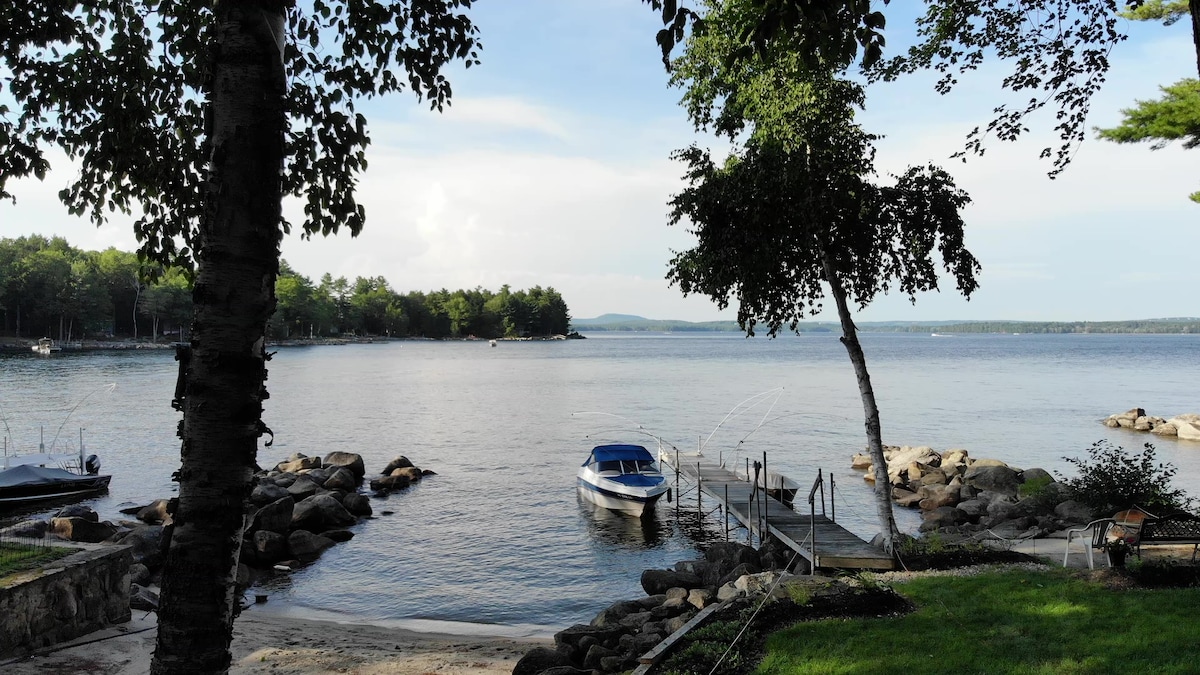 A serene waterfront scene is presented, showcasing a calm lake framed by trees. In the foreground, a small dock extends into the water alongside a moored boat. Rocky shorelines feature boulders partially submerged, adding to the natural beauty of the landscape.