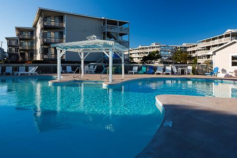 An inviting outdoor pool area features a spacious swimming pool, with a gazebo situated on a deck at the center. Surrounding lounge chairs provide ample seating, while the backdrop showcases several multi-story buildings of the resort, under a clear blue sky.