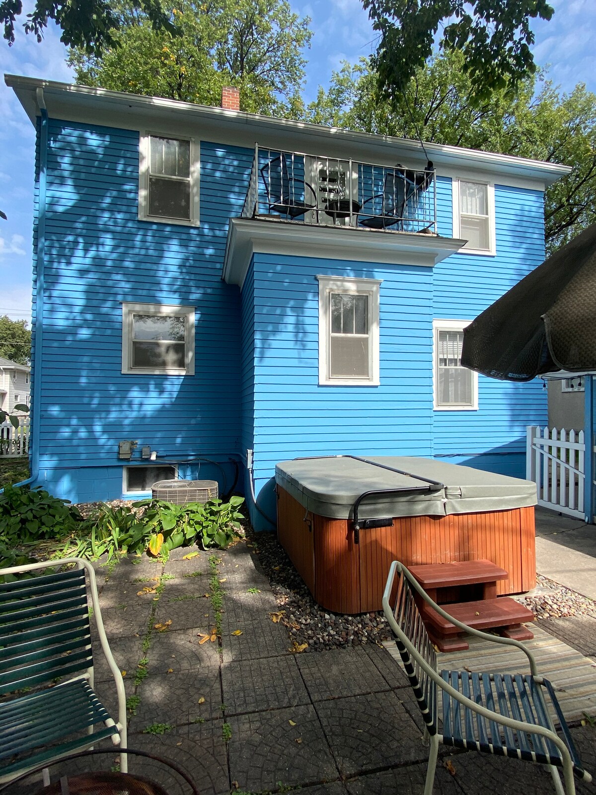 The exterior of a two-story blue house is displayed, featuring a balcony with black railings. A wooden hot tub is positioned on a paved area, surrounded by greenery and a few outdoor chairs. The property is enclosed by a white fence, providing a private atmosphere.