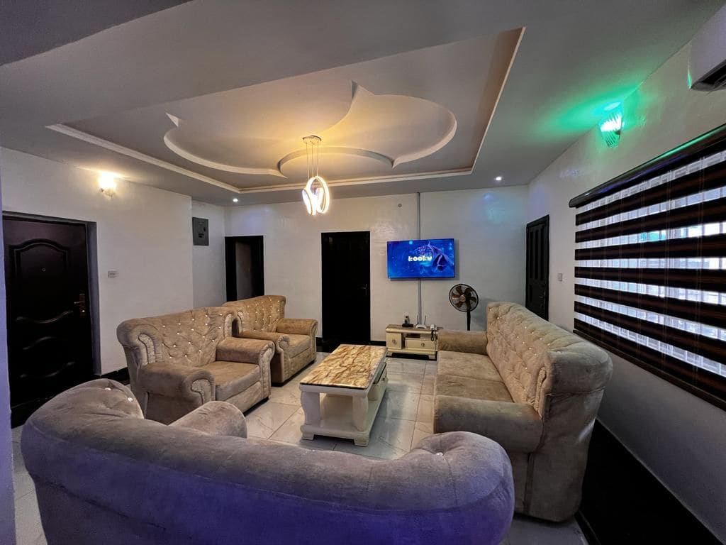 A spacious living area features a light-colored, plush seating arrangement, including two large sofas and matching armchairs. A modern coffee table sits at the center, and a wall-mounted TV is displayed prominently. Soft lighting creates a comfortable ambiance, complemented by stylish window treatments.