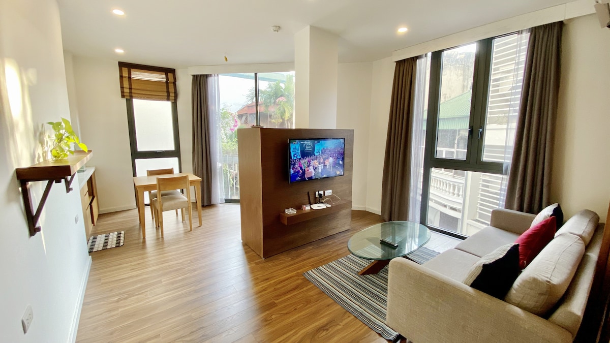 A modern living space features a comfortable sofa, a sleek glass coffee table, and a wall-mounted television. Natural light enters through large windows adorned with dark curtains. A small dining area is visible, complete with a wooden table and chairs, creating a functional yet cozy environment.