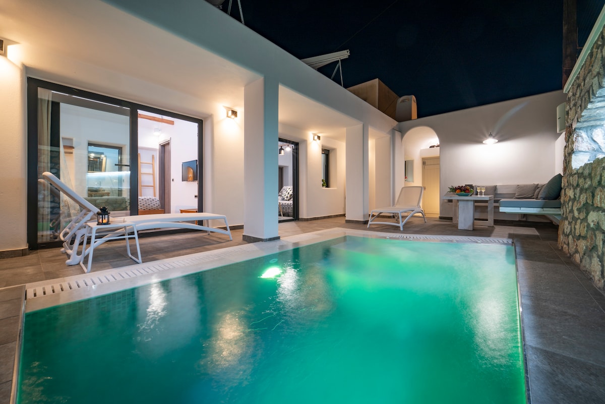 A private heated pool with a jacuzzi is centered in the outdoor area, surrounded by modern seating and loungers. Glass doors provide a view into the living space, reflecting soft interior lighting that enhances the serene ambiance. The layout promotes an inviting connection between indoor and outdoor settings.