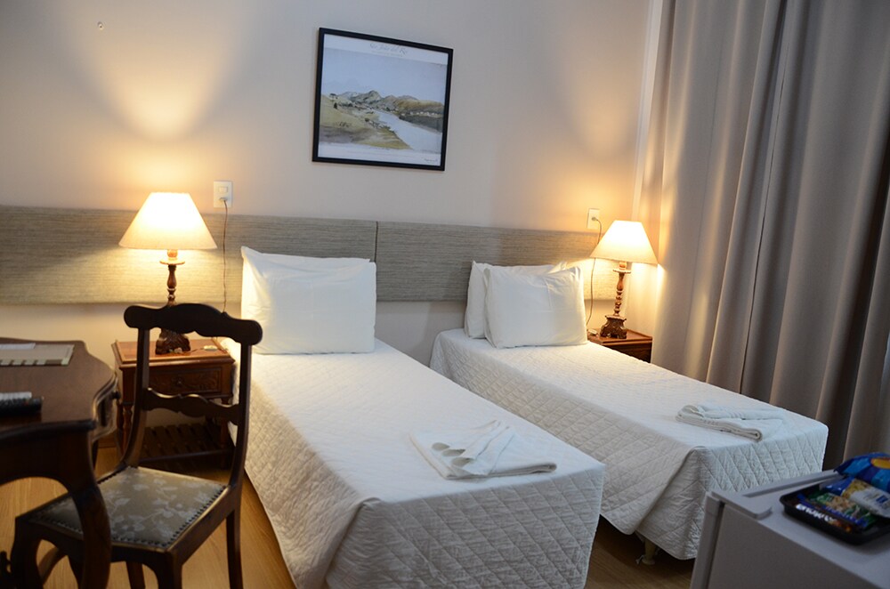The room features two spacious beds with white coverings, positioned beside wooden nightstands with lamps. A framed landscape artwork is hung on the wall above the beds. A wooden desk with a decorative chair is placed in the corner, with a small refrigerator visible to the side.