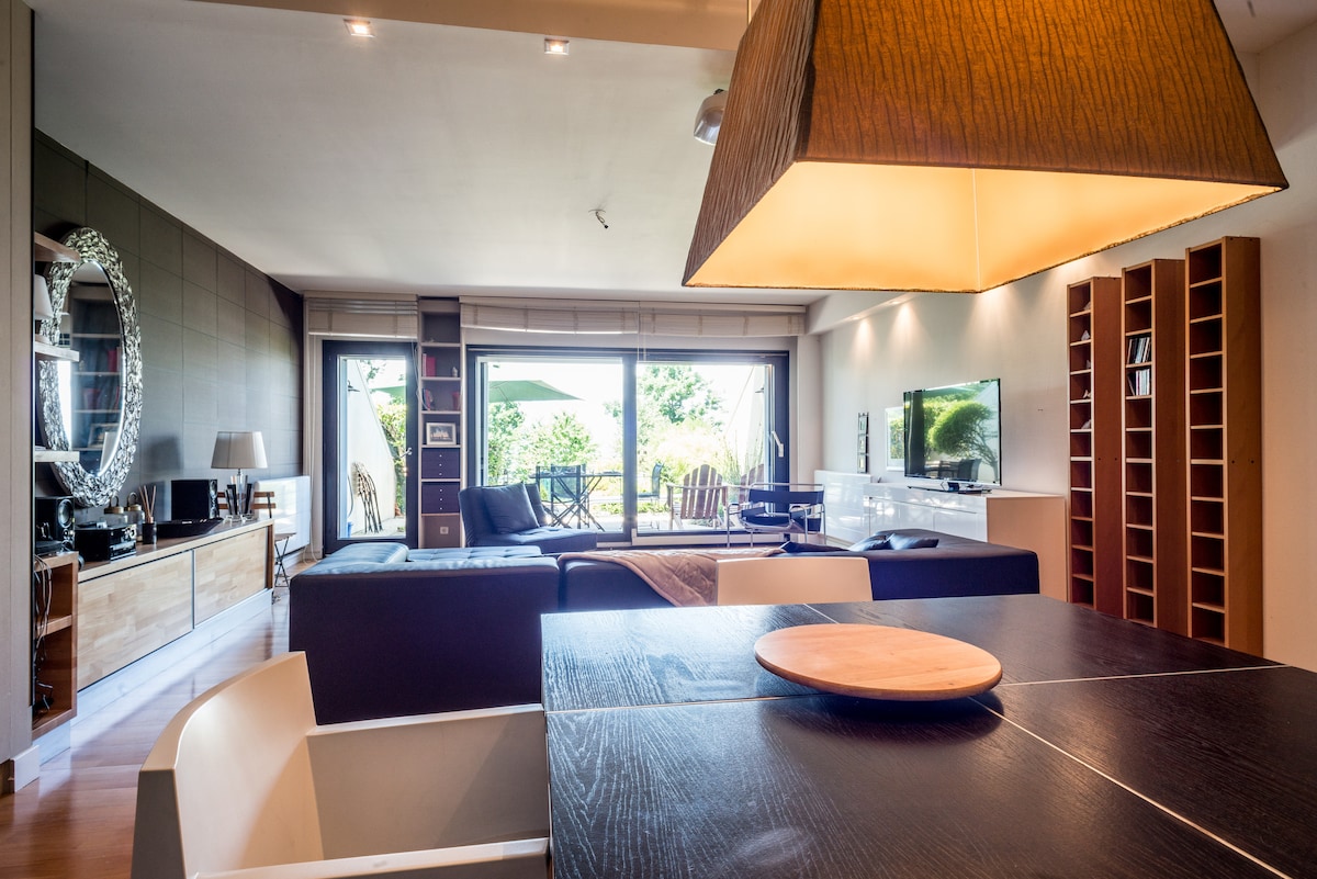 The open living area features large windows that provide views of the outdoors. A modern dining table is positioned in the foreground, with minimalist decor and a unique pendant lamp overhead. A comfortable seating arrangement is visible, along with various entertainment options on a wall-mounted television.