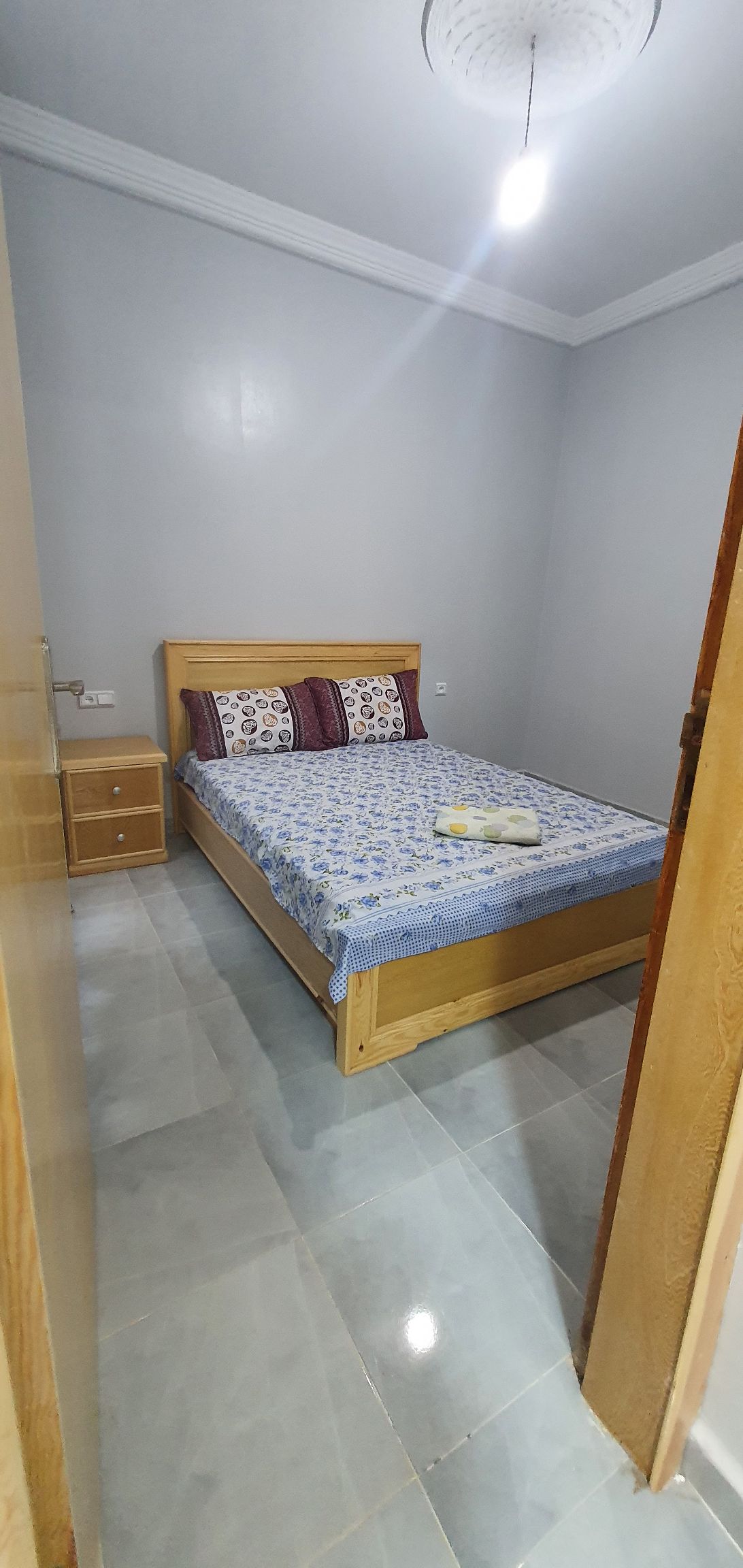 A cozy bedroom features a wooden bed with a floral patterned blanket and two decorative pillows. A bedside table is positioned nearby, and the room is illuminated by overhead lighting. The walls are painted in a soft gray, and a smooth tiled floor adds to the ambiance.