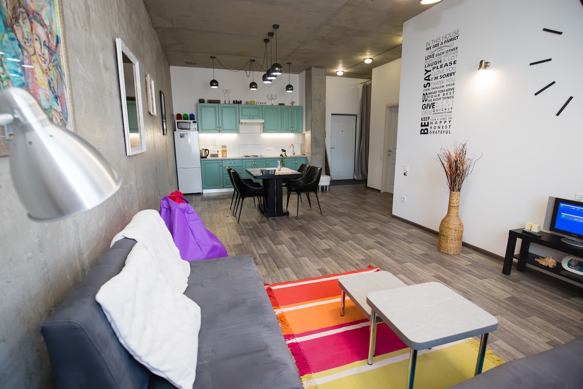 An open living area showcases contemporary design elements, featuring a comfortable gray sofa and vibrant area rug. A modern dining table with black chairs is positioned near a well-equipped kitchen. Soft lighting fixtures add a warm ambiance to the spacious environment.