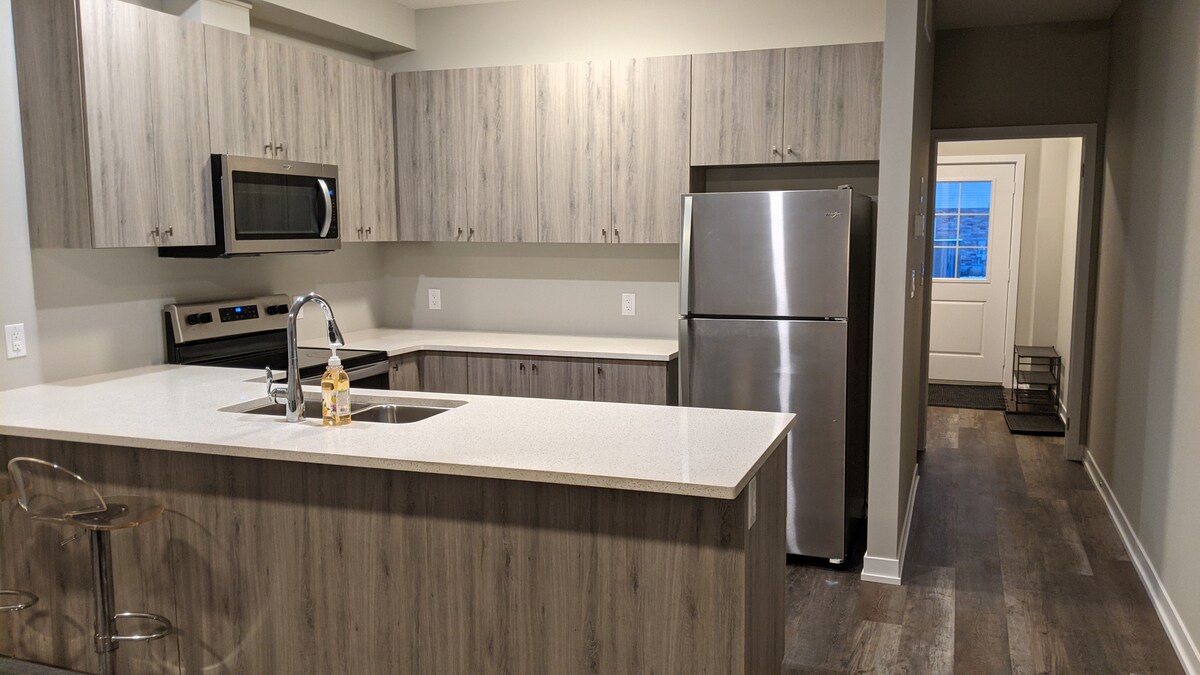 A modern kitchen features wood-grain cabinetry and stainless steel appliances, including a refrigerator and oven. A spacious countertop provides room for meal preparation, while a small decorative lantern adds a touch of warmth. The open layout connects to a neutral-toned hallway leading to the entrance.