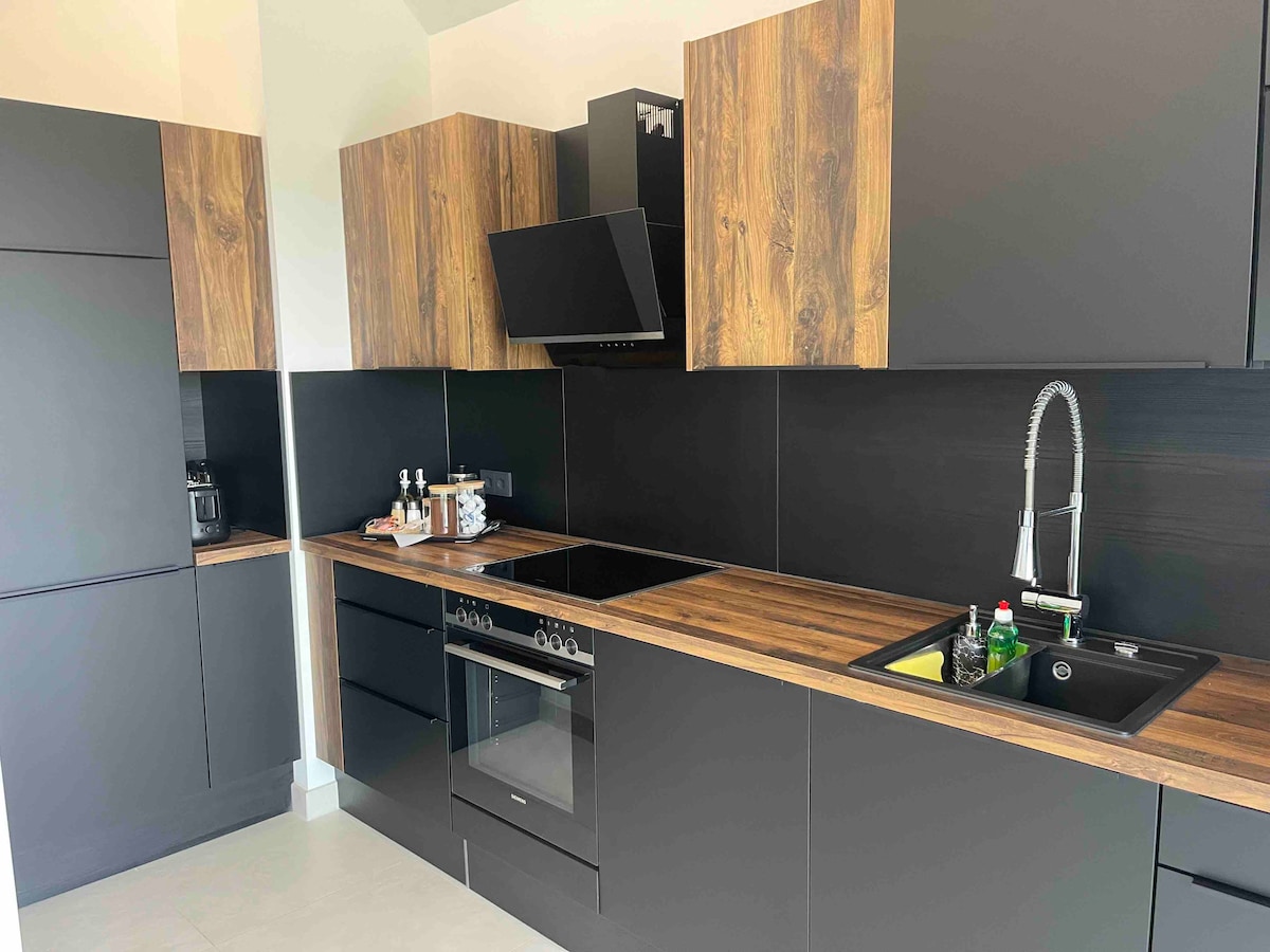 A modern kitchen is presented, featuring dark cabinetry combined with warm wood accents. A sleek stove is complemented by a black hood, while a sink with a polished faucet is positioned next to a cutting board. Kitchen essentials and small appliances are neatly arranged on the countertops.