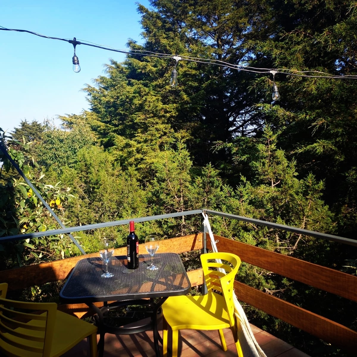 A small terrace is set among lush greenery, featuring a table with glasses and a wine bottle. Yellow chairs surround the table, while twinkling lights are strung overhead, enhancing the natural ambiance of the space.