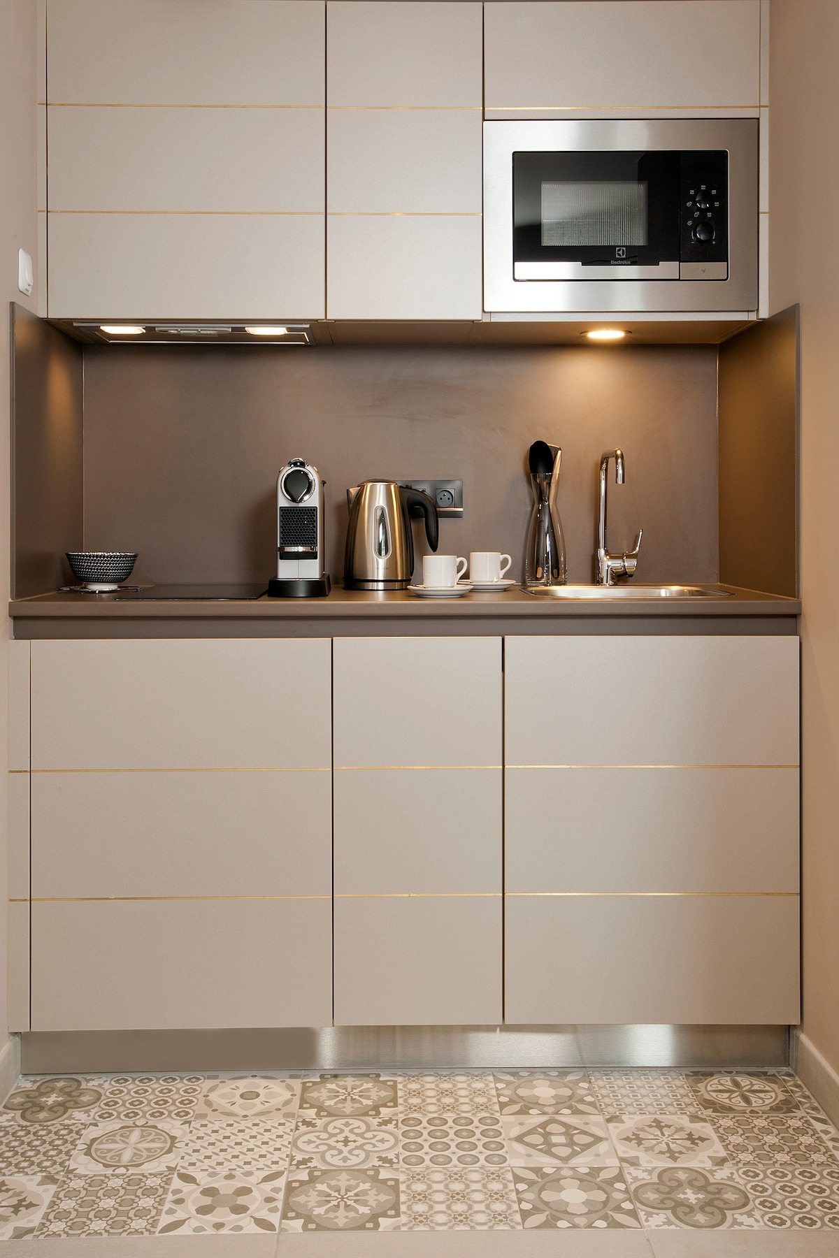 A modern kitchen area is highlighted with sleek cabinetry and a stylish countertop. An espresso machine and kettle are positioned next to two coffee cups. A microwave is mounted above, with under-cabinet lighting accentuating the design. Decorative patterned tiles cover the floor.