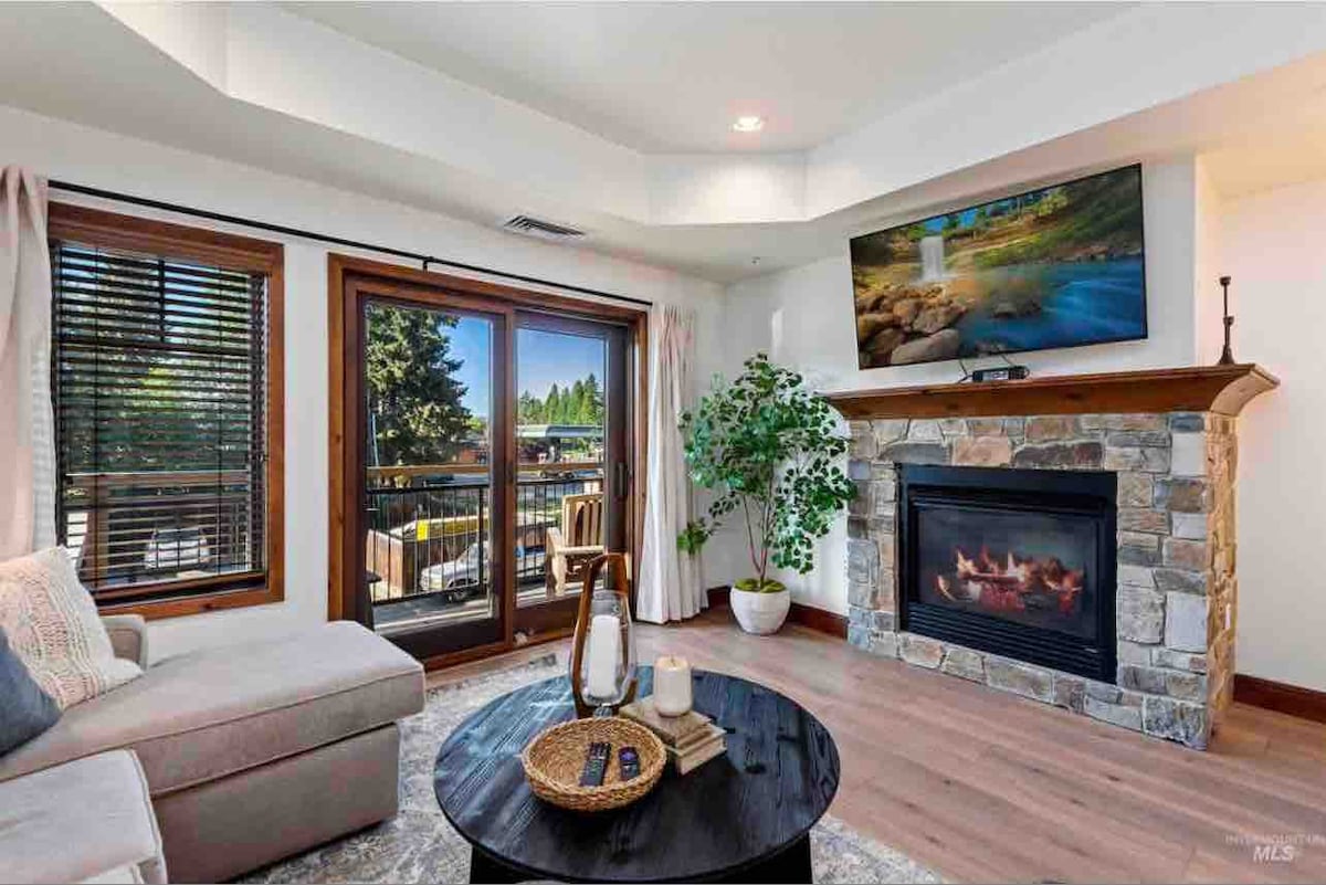 The living room features a comfortable sectional sofa positioned near a stone fireplace. A large flat-screen TV is mounted above the mantel. Natural light enters through double doors leading to a balcony, complemented by indoor plants and a round coffee table with decorative items.