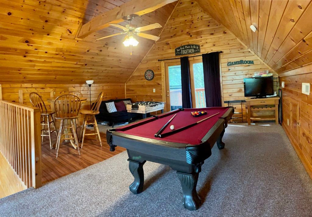 A game room features a pool table at its center, surrounded by wooden walls and a cozy ambiance. A plush couch and a table with chairs provide seating options. Natural light is let in through glass doors leading to a balcony, enhancing the inviting space.