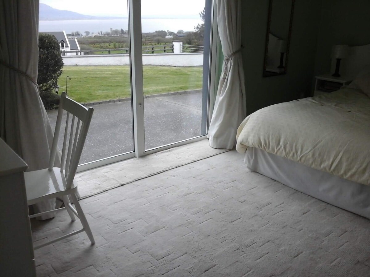 A bright bedroom features a spacious bed with neutral bedding. A white chair is positioned beside a glass door that opens to a view of the outdoor landscape. The floor is covered with a light-colored carpet, contributing to the room's airy feel.