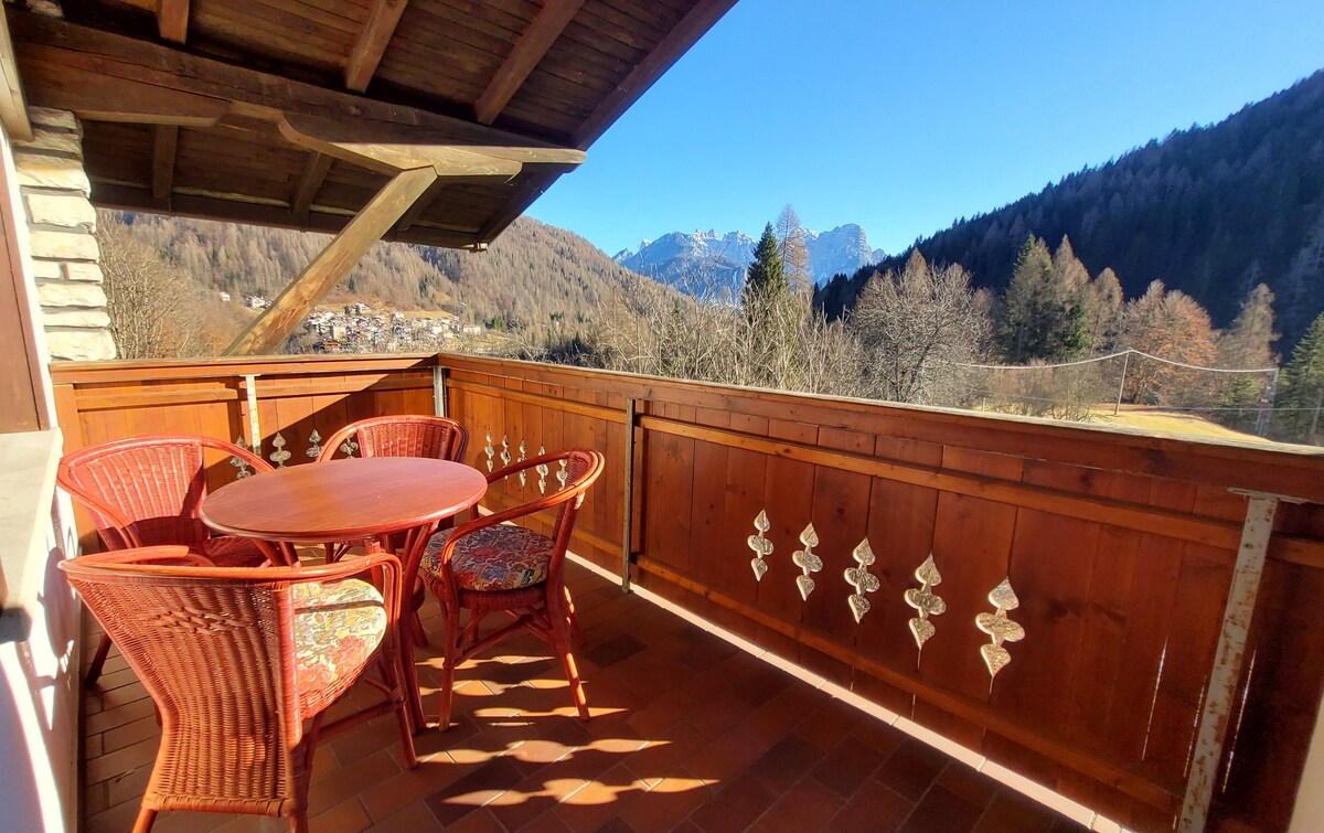 A spacious balcony features a round table surrounded by four woven chairs, adorned with floral cushions. Wooden paneling reflects traditional design elements, while panoramic views of the mountains and trees can be seen in the background. Sunlight casts a warm glow across the space.