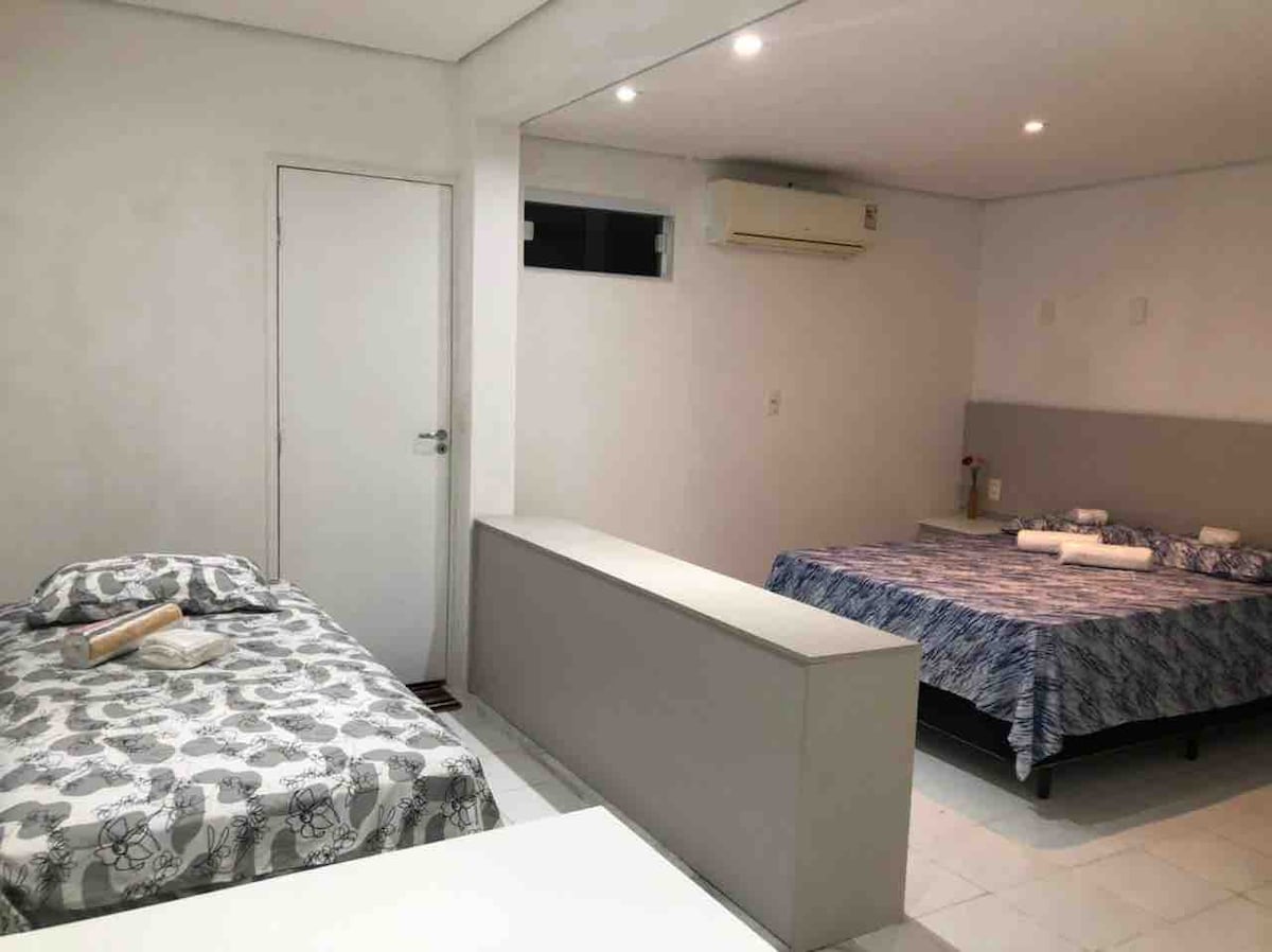A spacious studio room is presented, featuring two beds, one with a patterned duvet and the other with a solid blue cover. An air conditioning unit is mounted on the wall, and light fixtures illuminate the area. A door leads to an adjacent space.