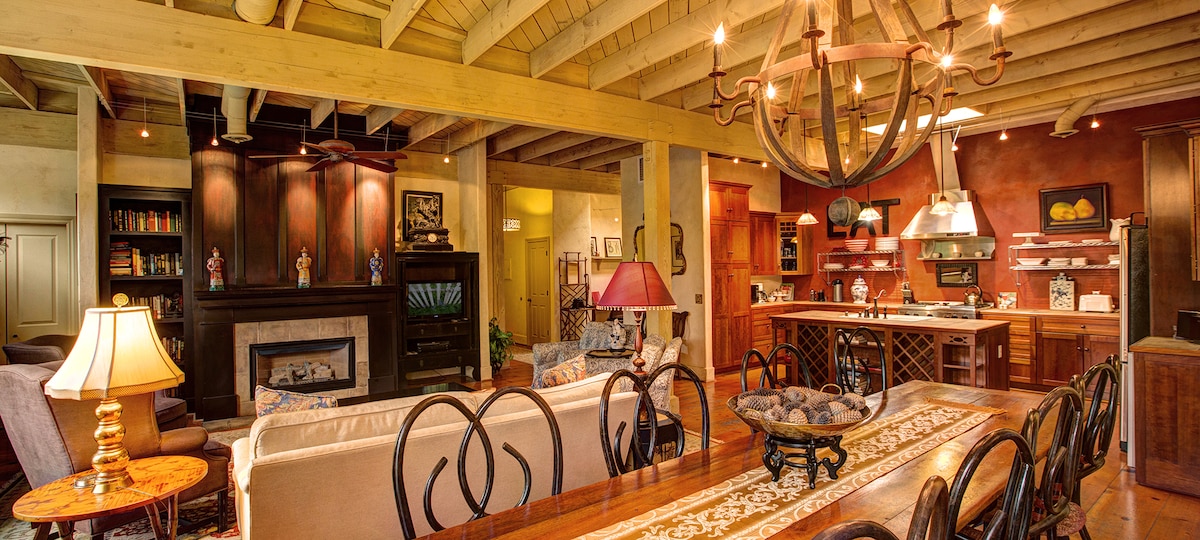 An expansive living area features a comfortable seating arrangement, including a sofa and armchairs, with a gas fireplace in the background. A large dining table is set in the foreground, surrounded by elegant chairs. The well-equipped kitchen is visible, showcasing modern appliances and ample counter space.