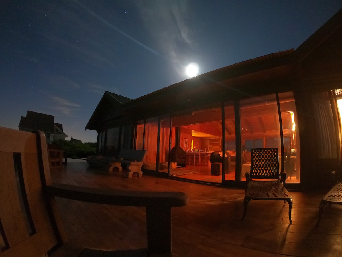 A wooden deck is illuminated by moonlight, featuring a variety of outdoor seating options. Large glass doors reveal the warm ambiance of the interior space, with a glimpse of the open plan living area. The night sky adds a serene backdrop to the scene.