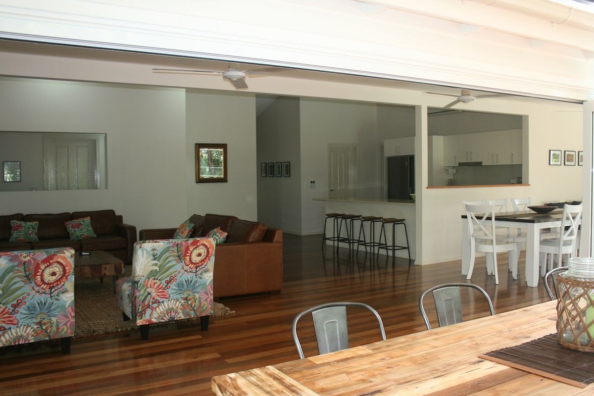 An open-plan living space is showcased, highlighting polished timber floors and natural light from large sliding doors. A dining area with a wooden table and metal chairs is visible, along with a cozy lounge area featuring upholstered seating and decorative cushions.