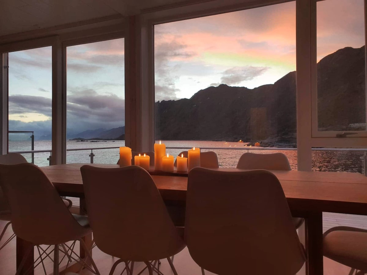 A dining area is visible with a wooden table and several white chairs arranged around it. Flickering candles are placed at the center, creating a serene ambiance, while large windows frame a stunning view of the ocean and mountains, enhanced by a colorful sunset.