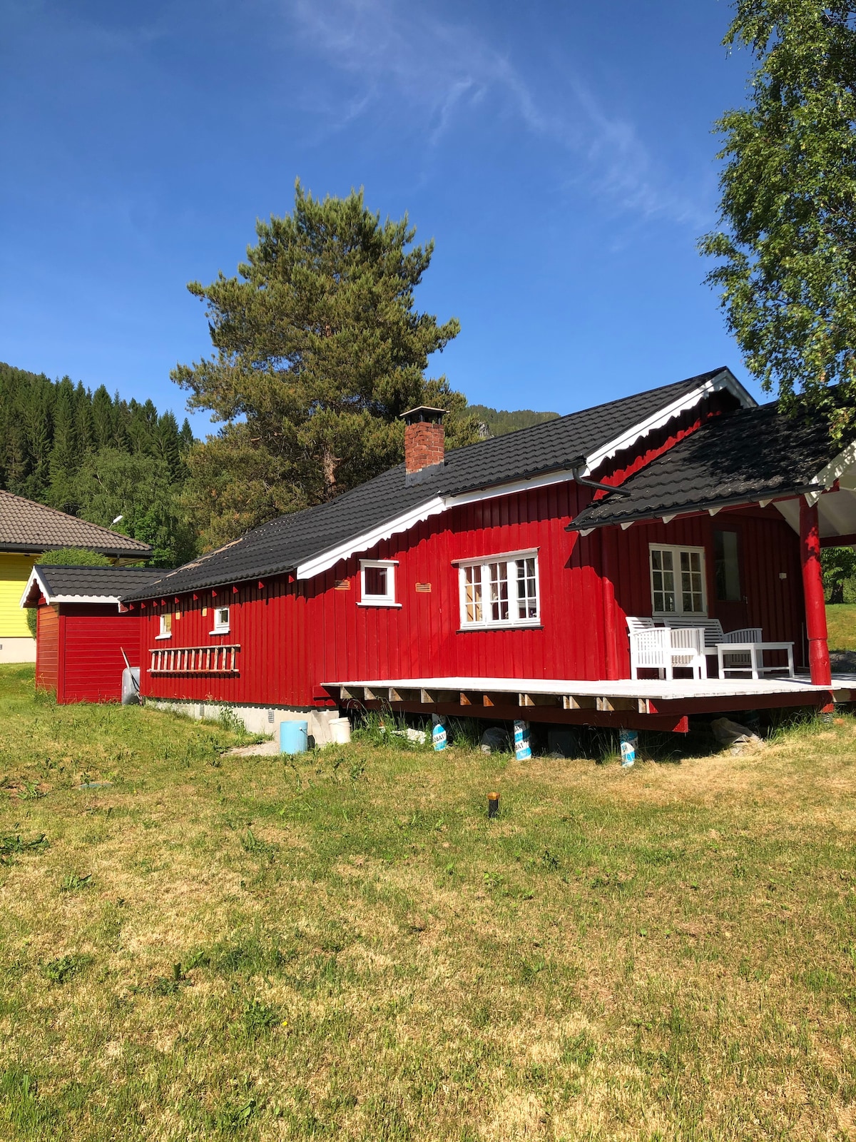 A charming red wooden cabin is set against a backdrop of greenery and blue sky. The structure features a black roof with decorative white trim, and a small porch with two white chairs. Lush grass surrounds the cabin, offering a tranquil outdoor space.