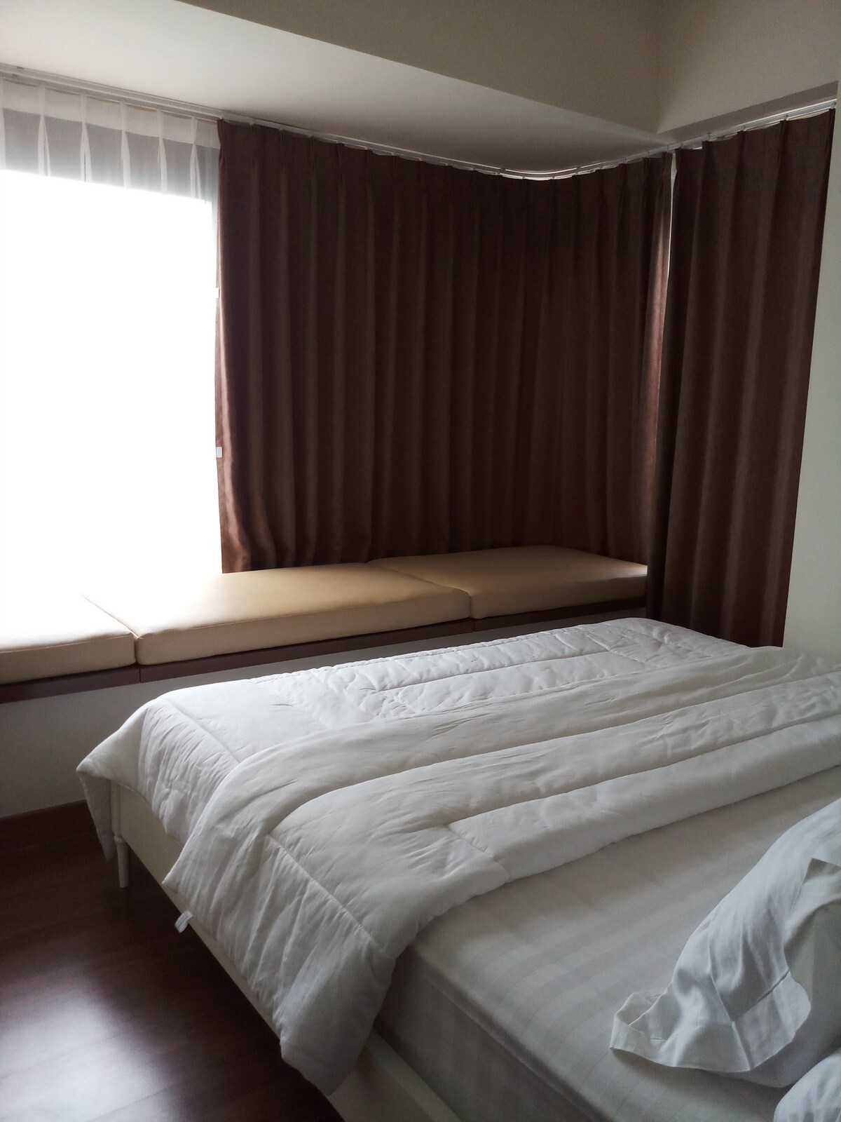 A cozy sleeping area features a spacious bed with a white comforter and a neatly arranged blanket. Natural light enters through a large window, complemented by dark brown curtains. A cushioned nook by the window provides a comfortable place to relax.