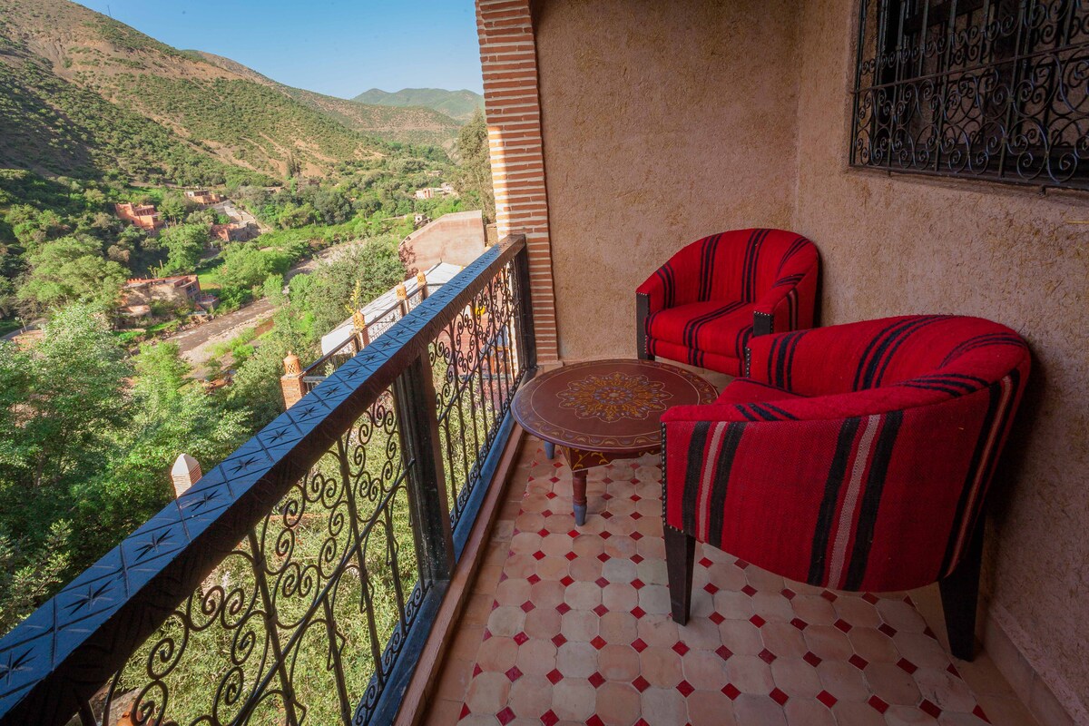A private terrace features two red patterned chairs and a circular table, offering a view of the surrounding mountains and greenery. The floor is adorned with decorative tile work, and a wrought-iron railing frames the space.