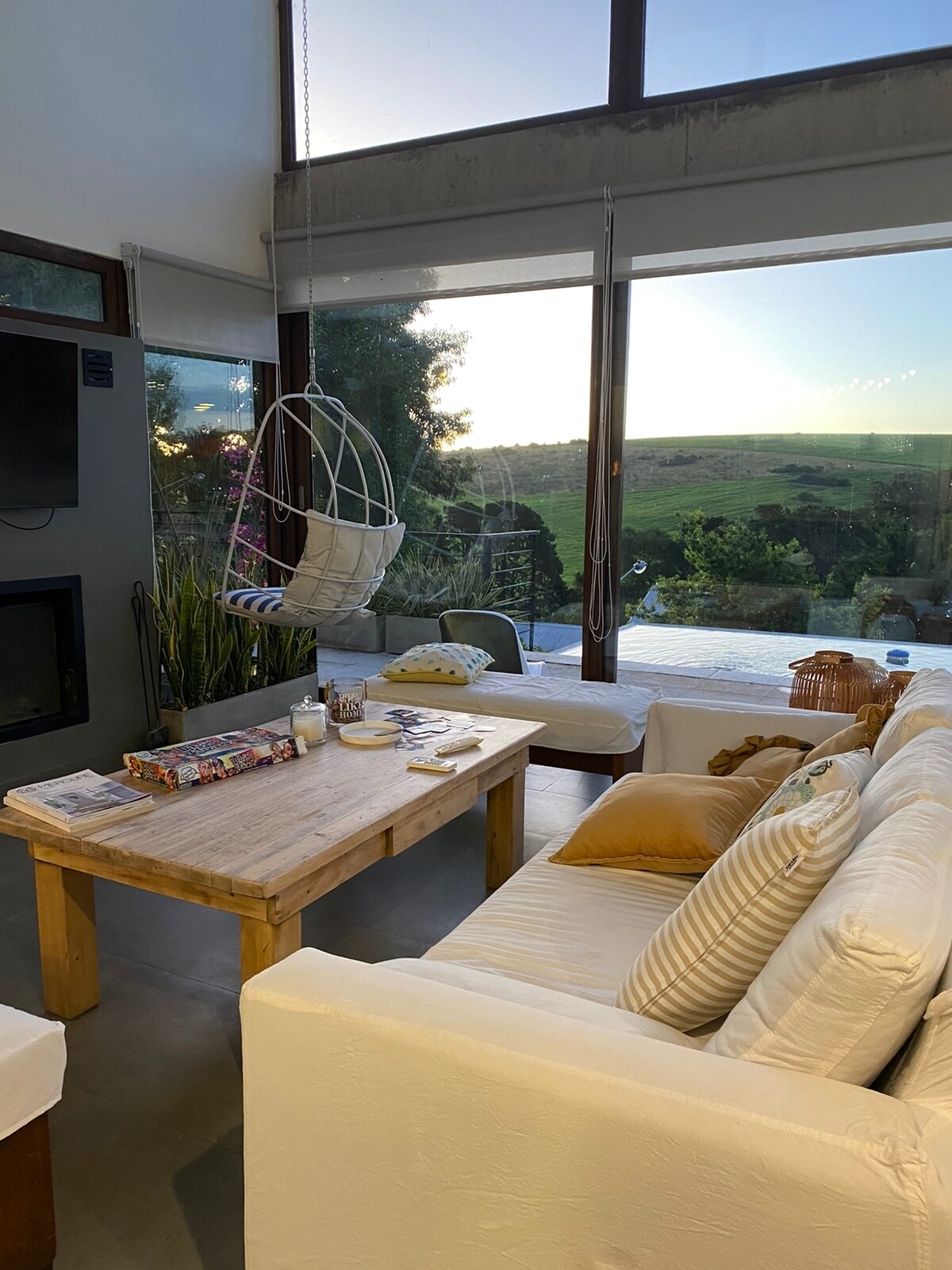 A comfortable seating area features a white sofa and a rustic wooden table adorned with various books and candles. A hanging chair sways gently, while large glass windows frame scenic views of the rolling landscapes and a serene outdoor space.