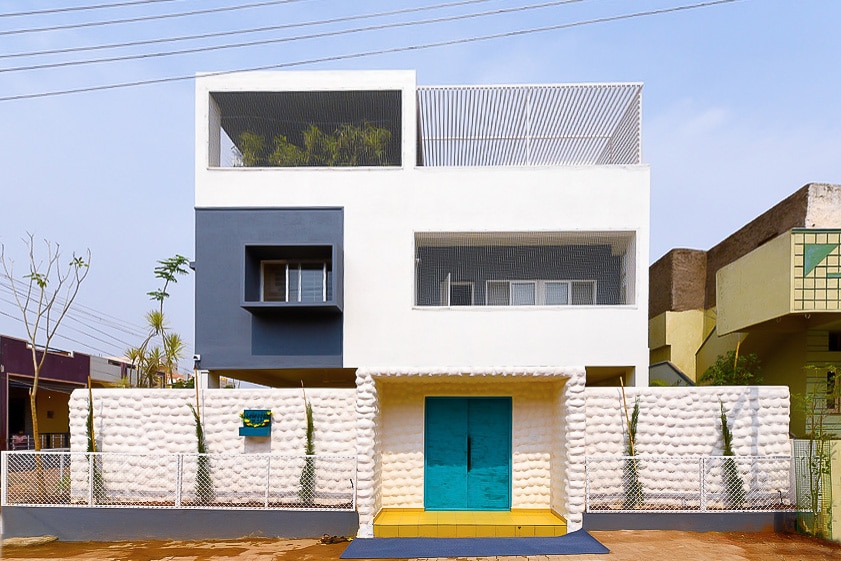 The exterior of the villa features a modern architectural design with a bold turquoise door and distinct blue and white color blocks. Decorative elements include a textured wall and landscaped plants, contributing to a contemporary aesthetic. The second floor is highlighted by an open terrace surrounded by railings.