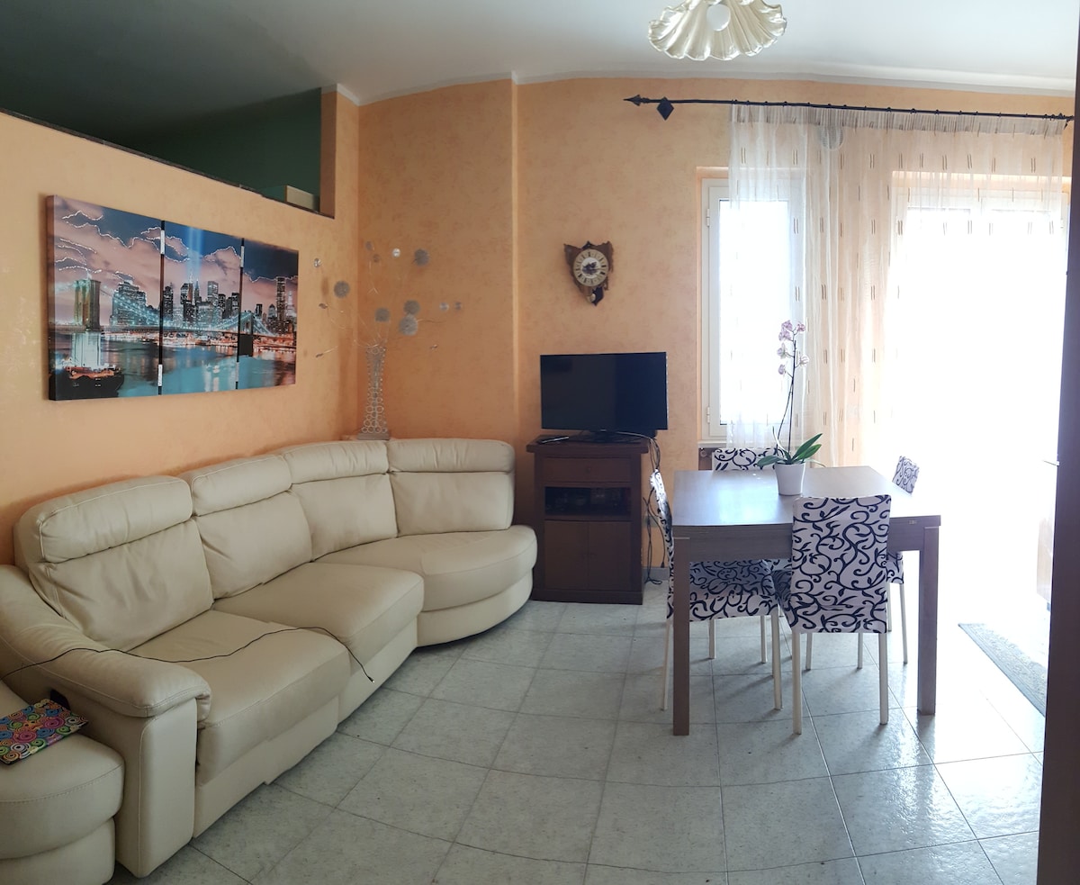 A living area features a light-colored sectional sofa and a wooden dining table surrounded by patterned chairs. A television rests on a stand, and natural light enters through large windows with sheer curtains. A decorative wall piece adds interest to the space.